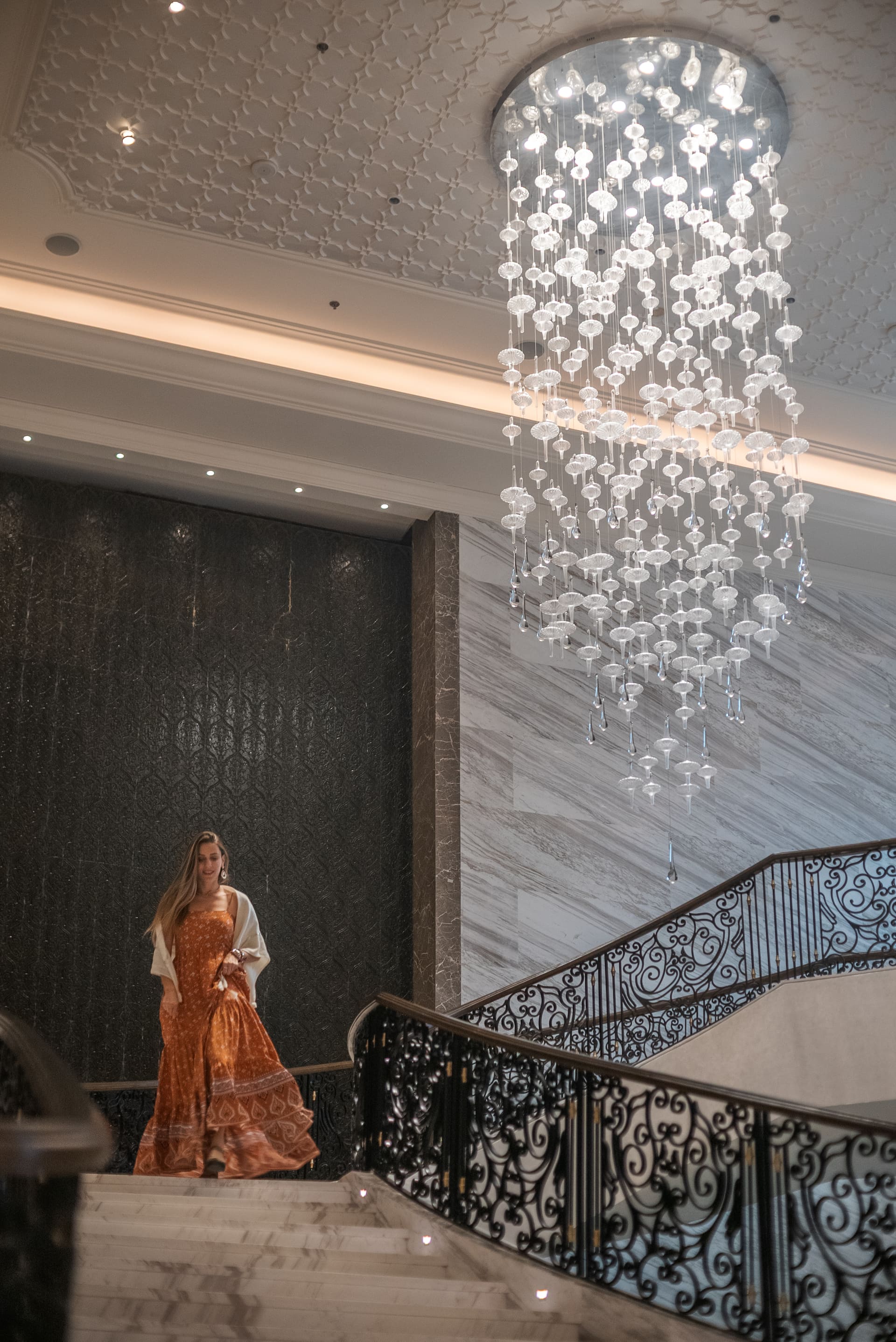 Mulher de vestido longo alaranjado descendo a escadaria de marmore de um hotel, sob um lustre de cristais.