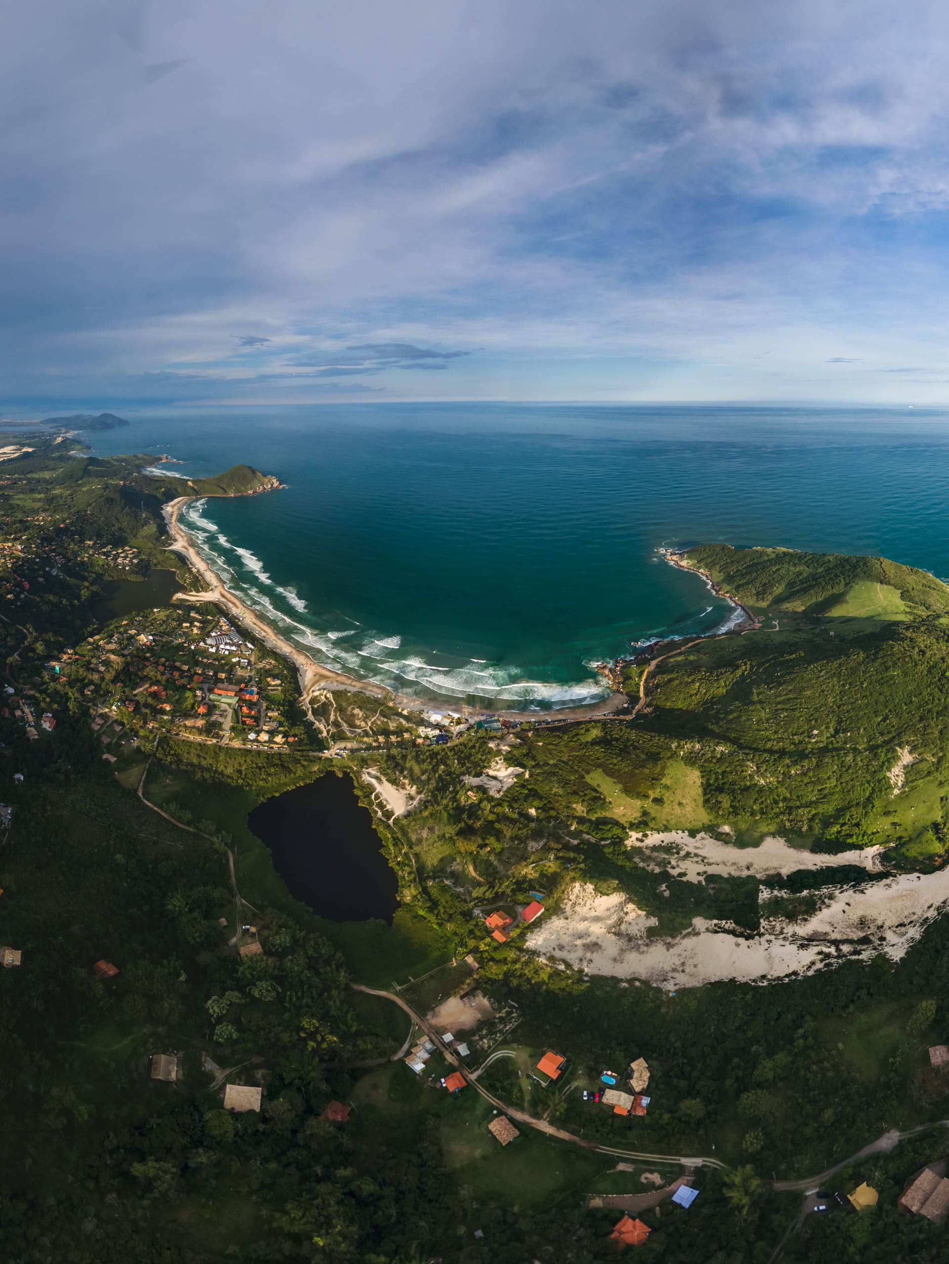 Vista aerea da faixa litoranea: praia, lagoa escura, dunas e casario na mata.