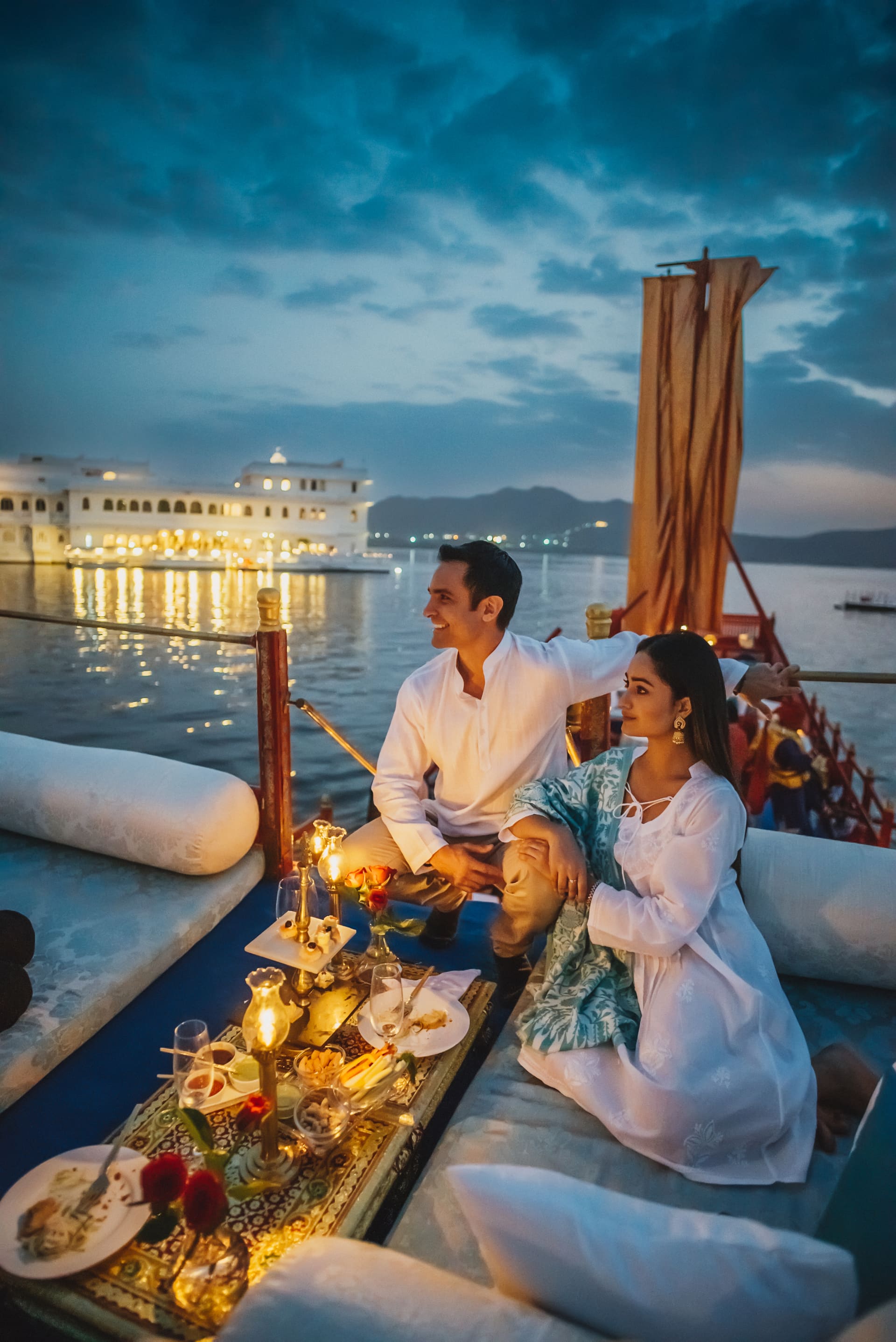 Casal vestido de branco a beira do lago ao por do sol, palacio aceso na agua.