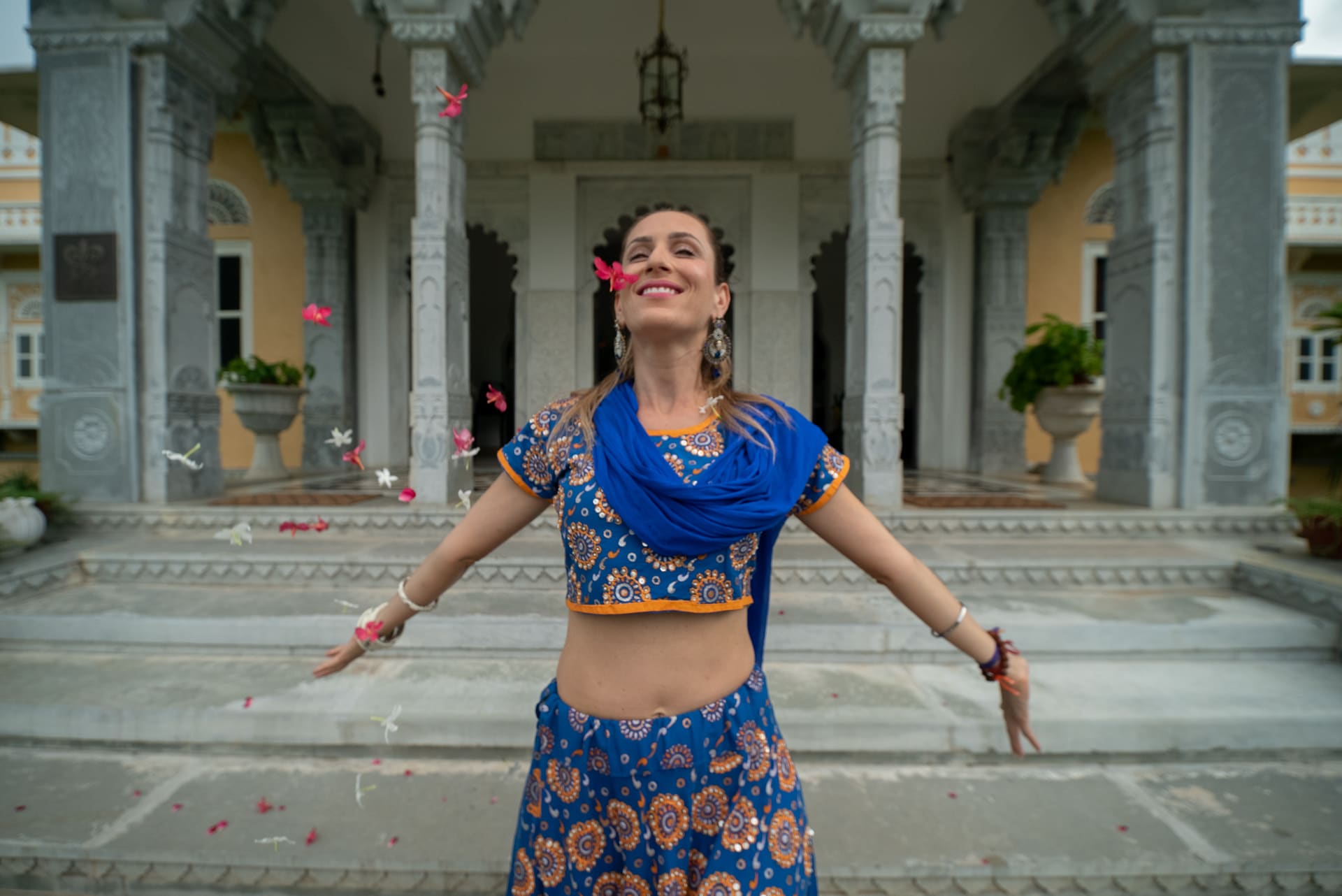 Mulher de traje azul bordado, de bracos abertos entre petalas, diante da colunata de um palacio.