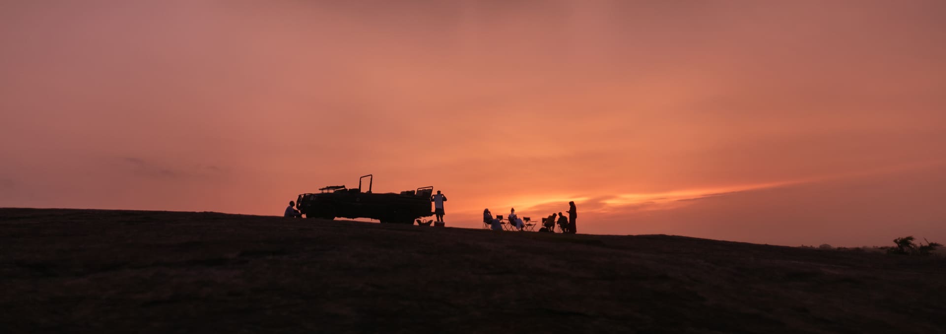 Silhuetas de um jipe e de um grupo de pessoas na crista de um morro, contra o ceu alaranjado do por do sol.