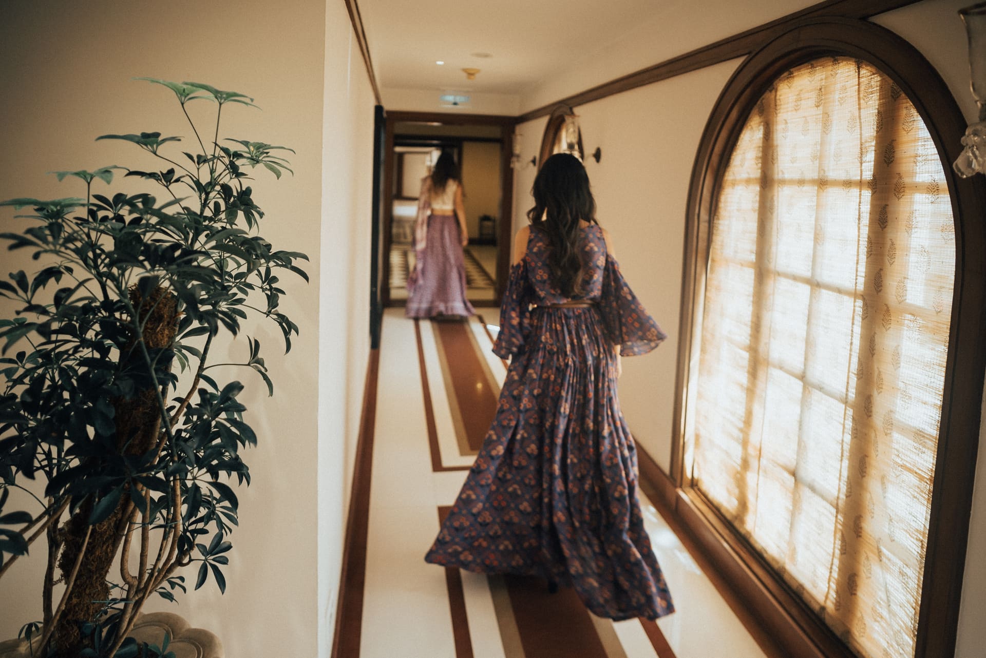 Duas mulheres de vestido longo num corredor de hotel, uma refletida no espelho, luz da janela em arco.